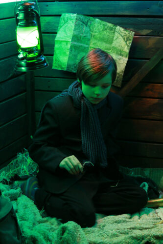 Ein Junge sitzt mit Kissen und Decke und einer Lampe in einem abgedunkelten Raum.