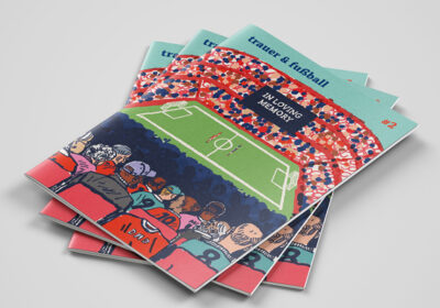 Das Titelbild des Fanzines "trauer & fußball" #2 ist eine Illustration: Ein volles Stadion. Auf dem schwarzen Big Screen steht in weißer Schrift "In loving memory". Auf dem Rasen halten beide Mannschaften am Mittelkreis eine Gedenkminute ab. Von hinten sieht man diverse Fans auf Sitzen.